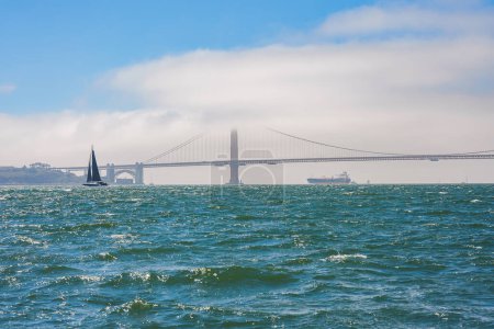 Foto de Vista serena de la bahía de San Francisco con el emblemático puente Golden Gate en el fondo. Niebla, velero, buque de carga y aguas tranquilas crean una hermosa escena. - Imagen libre de derechos