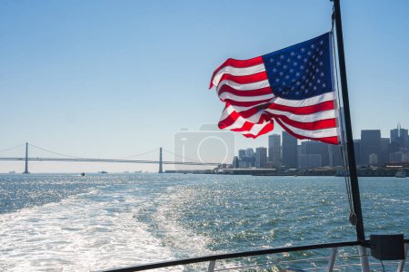 American flag fluttering on boat as it leaves SF towards Bay Bridge. Skyline, buildings, river wake visible on sunny day. Maritime culture essence captured.