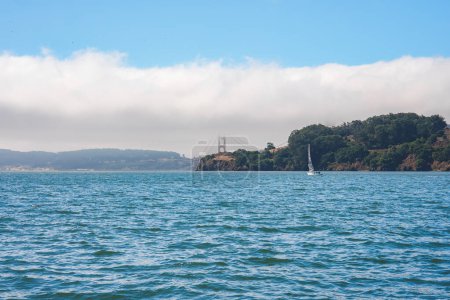 Gelassener Blick auf die Bucht von San Francisco mit teilweise verhüllter Golden Gate Bridge im Nebel. Blaues Wasser, weißes Segelboot, grüne Landmasse und wolkenverhangener Himmel tragen zu ruhiger Schönheit bei. Ideal für Reiseblogs.