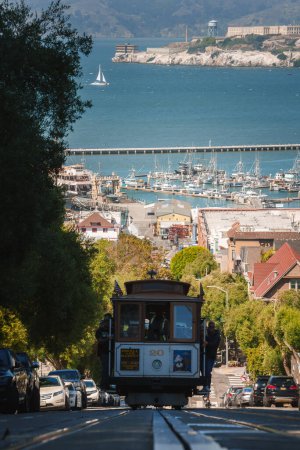 Photo for Iconic San Francisco cable car ascending steep hill with passengers enjoying the ride. Marina boats, Alcatraz Island in distance. Rich history and maritime culture captured. - Royalty Free Image