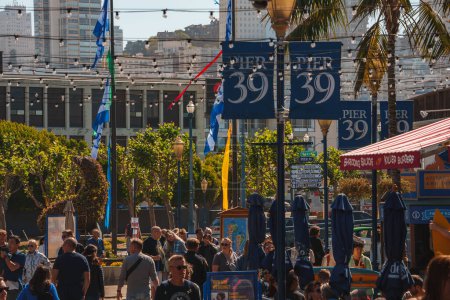 Photo for Bustling scene at Pier 39, San Francisco. Focus on blue and gold signs above crowd, lively atmosphere with visitors, palm trees, city architecture, flags, sunny day vibe. - Royalty Free Image