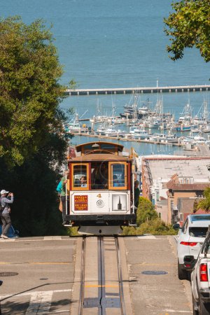 Photo for Classic San Francisco cable car ascending steep hill, wooden with brown and cream color scheme. Marina with boats in background, near Fishermans Wharf. - Royalty Free Image