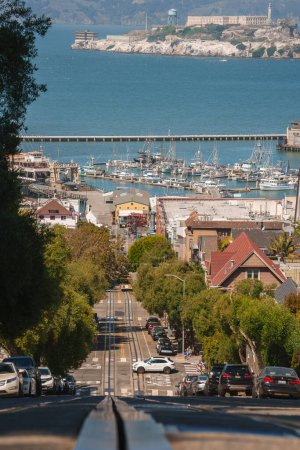 Photo for View looking down a steep San Francisco street towards a marina filled with yachts and boats, including Alcatraz Island in the distance. Parked cars line the street with sunny skies. - Royalty Free Image