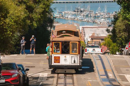 Photo for Iconic cable car travels down a steep San Francisco street, tracks visible, bay in background with boats, tourists take photos, urban nature scene with clear skies, bright day. - Royalty Free Image