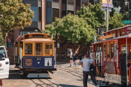 Photo for Vibrant street scene showcases iconic San Francisco cable cars with operator in white shirt and dark pants. City area setting with pedestrians and historic charm. Sunny day atmosphere. - Royalty Free Image