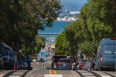 Photo for View down a steep street in San Francisco lined with cars. Cable car tracks visible. Cityscape with bay, boats, and blue sky. Urban life scene near Pier 39. - Royalty Free Image