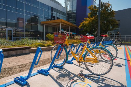 Photo for Bright yellow bicycle with red basket in blue bike rack on paved area with painted lines. Background shows modern building, sunny weather, lush greenery. - Royalty Free Image