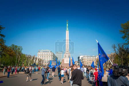 Téléchargez les photos : Rassemblement dynamique dans la vieille ville de Riga, Lettonie. Personnes portant des drapeaux de l'UE et de la Lettonie sous un ciel bleu clair. Monument de la liberté en arrière-plan. Une atmosphère festive et paisible célèbre le 20e anniversaire de l'UE. - en image libre de droit