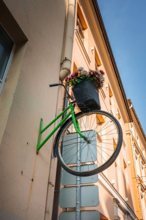 Instalación urbana con una bicicleta verde en una pared, adornada con flores de colores sobre cajas eléctricas grises. Posiblemente situado en el casco antiguo de Riga, bajo un cielo despejado.