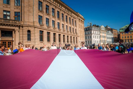 Foto de Escena vibrante en el casco antiguo de Rigas con una multitud celebrando bajo una gran bandera letona sobre un telón de fondo de cielo azul. Edificios históricos enmarcan el ambiente festivo de unidad y orgullo. - Imagen libre de derechos