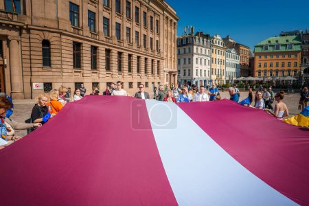 Foto de La escena de celebración en Riga, Letonia, muestra a una multitud diversa ondeando una gran bandera letona contra el telón de fondo del cielo azul. Ambiente festivo con banderas de la UE y arquitectura histórica. - Imagen libre de derechos