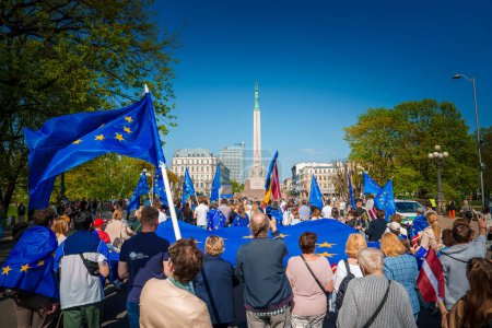 Téléchargez les photos : Rassemblement dynamique dans la vieille ville de Riga, en Lettonie, avec des drapeaux de l'UE et de la Lettonie célébrant le 20e anniversaire. Iconic Freedom Monument se dresse en arrière-plan. La foule unie dans la fierté. - en image libre de droit