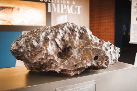 A large, irregular meteorite on display in a museum exhibit. Its rough, metallic surface shows signs of atmospheric entry. Text indicates its from Meteor Crater, Arizona, USA.