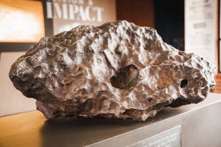Large, textured meteorite from Meteor Crater, Arizona, USA. Indentations and holes mark its high speed entry. Displayed on stand with educational panel background.