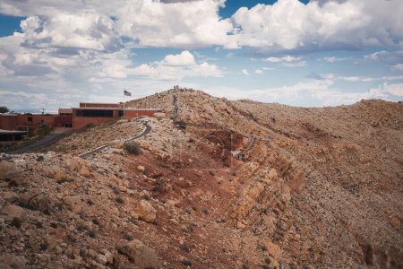 Descubra un terreno rocoso con un sendero sinuoso, una plataforma de observación y un centro de visitantes contra un cielo parcialmente nublado. Probablemente cerca de Meteor Crater, Arizona, EE.UU..