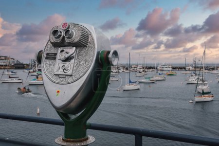Discover a coin operated binocular viewer on a railing overlooking a harbor filled with boats at a coastal marina. Beautiful sunset sky in the background.