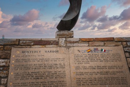 Historische Gedenktafel an einer Steinmauer mit Metallskulpturen, Fahnen und Leuchtturm in der Nähe des Hafens von Monterey. Ausführlicher Text auf Gedenktafel über maritime Bedeutung. Himmel mit warmen Farben des Sonnenuntergangs.