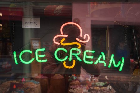 Photo for Neon sign featuring ICE CREAM in vibrant green letters, with orange and white ice cream cone icon. Placed on storefront window with visible items, daytime reflection. - Royalty Free Image
