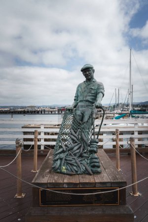 Fishing statue of a traditional fisherman on a pier with boats in the background. Serene maritime scene with cloudy sky. Ideal for coastal community concepts.