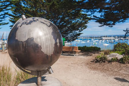 Photo for Metallic globe sculpture on stand with reflective surface featuring continents outlined against clear blue sky. Harbor view with boats and lush greenery in background. Peaceful Monterey, USA. - Royalty Free Image