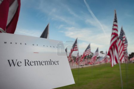 Close up view of sign saying WAVES OF FLAGS with We Remember in larger font, underlined in red. Background has American flags in California, memorial.