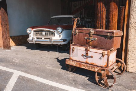 Photo for Vintage scene in Barstow, USA along Route 66. Rusted luggage cart with leather suitcases, classic car from 1950s 60s, sunny day evoking Americana nostalgia. - Royalty Free Image