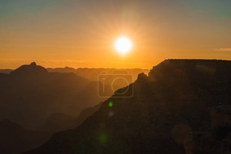 Téléchargez les photos : Scène de coucher de soleil serein avec le soleil bas dans le ciel jetant une lueur dorée chaude. Les crêtes de montagne créent une silhouette contre le ciel dans une image atmosphérique. - en image libre de droit
