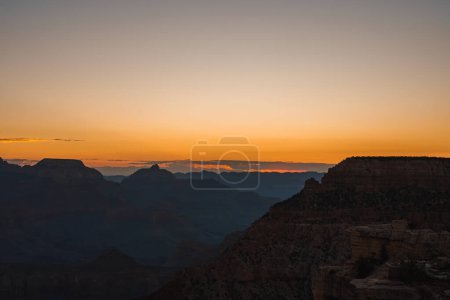 Szenischer Sonnenuntergang über einer riesigen Schlucht mit warmen Farbverläufen von Orange bis Blau. Silhouetten von Felsformationen, die gegen den Abendhimmel sichtbar sind. Möglicherweise Aussichtspunkt Grand Canyon. Atemberaubende natürliche Schönheit.