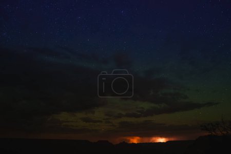 Ciel nocturne avec étoiles sur paysage sombre, silhouettes de mesas contre ciel plus clair. Nuages et lueur brillante visibles au Grand Canyon.