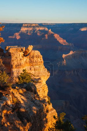Stunning view of Grand Canyon with distinctive geological formations, warm golden light at sunrise or sunset, clear blue sky, deep gorges carved by Colorado River. Location likely South Rim.