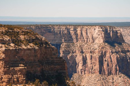 Vaste canyon avec des falaises escarpées et des formations rocheuses stratifiées dans des tons de rouge, brun et beige. Végétation clairsemée et ciel bleu clair. Ressemble aux paysages du sud-ouest américain.