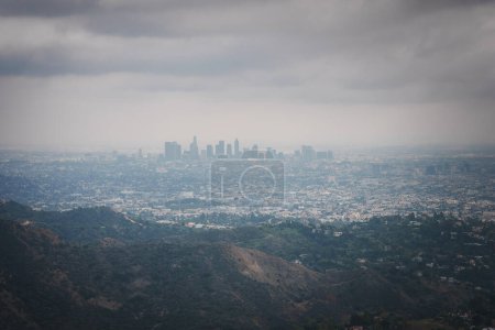 Vue aérienne d'un vaste paysage urbain sous un ciel nuageux, ligne d'horizon du centre-ville de LA au loin. Collines ondulantes au premier plan, probablement Hollywood Hills, bâtiments denses avec un éclairage doux. Capture de point de vue élevé.