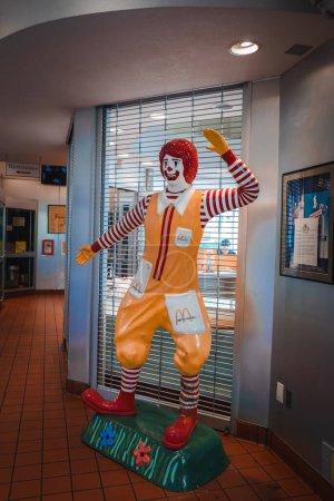 Téléchargez les photos : Statue Ronald McDonald grandeur nature en tenue iconique, agitant et accueillant. Intérieur avec stores, probablement à Los Angeles. Conseils de fond au cadre du restaurant. - en image libre de droit