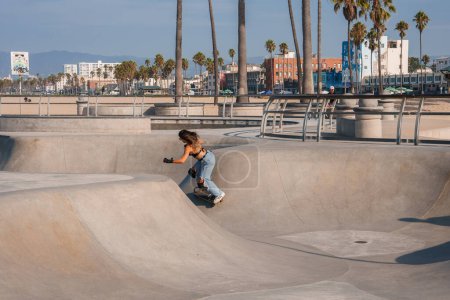 Photo for Skateboarder performing tricks at Venice Beach Skatepark in Los Angeles, California. Woman with long hair crouched in concrete bowl, capturing sunny outdoor vibe. - Royalty Free Image