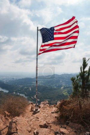 Photo for American flag on flagpole waving against hilly backdrop. Rugged terrain leads to flagpole, cloudy sky with natural light. View reminiscent of Los Angeles hills. - Royalty Free Image