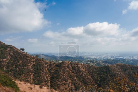 Téléchargez les photos : Vue panoramique sur les collines arides entourant Los Angeles avec un paysage urbain au loin sous un ciel partiellement nuageux. Hélicoptère ajoute du dynamisme à la scène tranquille. - en image libre de droit