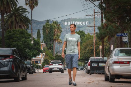 Photo for Man walking on residential street in LA with Hollywood sign in background, lush greenery, and parked cars. Casual attire, overcast sky. Hollywood Hills view. - Royalty Free Image