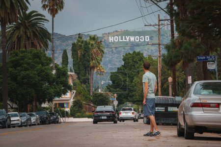 Photo for Urban scene in Los Angeles featuring a figure gazing at the Hollywood sign. Parked cars, palm trees, and gloomy sky in the background. Ideal for entertainment industry websites, blogs, or magazines. - Royalty Free Image