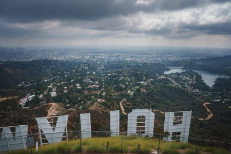 Photo for Iconic Hollywood Sign view over Los Angeles hills and cityscape under cloudy sky. Letters seen from behind on hilly, lush terrain with a body of water. - Royalty Free Image