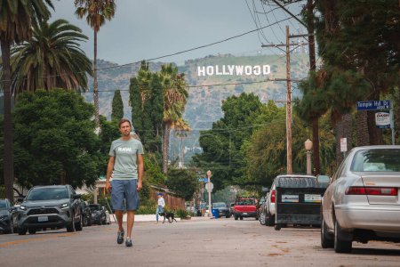 Photo for Man strolling down LA street with parked cars, palm trees. Hollywood sign visible, cloudy sky. Casual attire, relaxed gait. Hollywood neighborhood vibes. - Royalty Free Image
