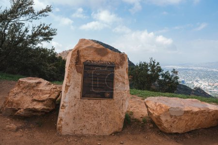 Large stone slab with bronze plaque surrounded by boulders. Panoramic city view in the background, possibly taken in hills near Los Angeles for reflection.