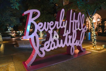 Photo for Nighttime scene in Beverly Hills with a decorative sign reading Beverly Hills, illuminated against lush greenery and elegant architecture. Serene and inviting ambiance. - Royalty Free Image
