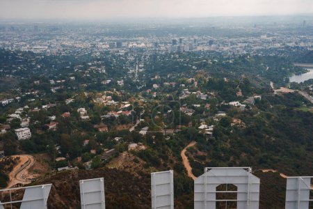 Iconic Hollywood Sign overlooking Los Angeles from elevated viewpoint in the hills. Greenery, urban development, winding roads, and cityscape in the distance.