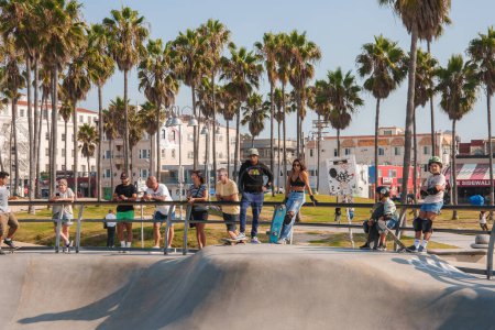 Photo for Sunny day at a skatepark in Venice Beach, Los Angeles. Skaters gathering in a concrete bowl surrounded by palm trees and city buildings. Casual atmosphere. - Royalty Free Image