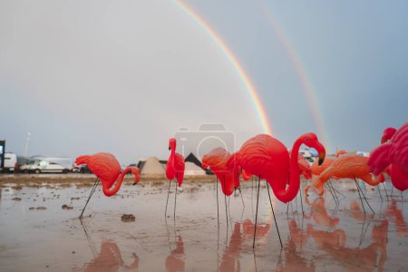 Lebendige rosa Flamingo-Ornamente in schlammiger Wüste unter einem doppelten Regenbogen. Surreale Szene mit Spiegelungen, Zelten, Fahrzeugen und Festival-Vibes.getLocale.