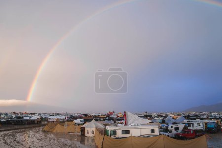 Sprawling desert settlement with wet ground and rainbow sky. Mix of tents, RVs, and structures. Transient city amidst barren landscape. Mountain range in distance.