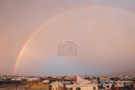 A music and art festival hosted in a desert, featuring a vibrant community living in tents and makeshift shelters, under a twilight sky with a rainbow.