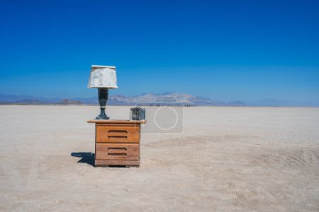 Vintage wooden dresser with lamp in desert landscape. Surreal setting with cracked ground, distant mountains, and blue sky. Art installation at music festival. Evokes abandonment and curiosity.