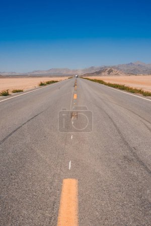 Route panoramique à travers un paysage aride avec un ciel bleu clair, des traces de pneus sur l'asphalte, menant vers des collines en Californie. Éloignement et tranquillité capturés.