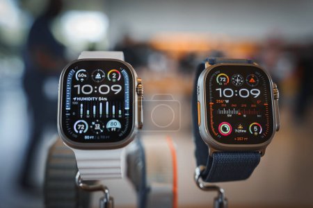 Photo for Two Apple Watches, one silver with white band, one dark with black loop, display the time as 10 09 in a blurry Apple Store background with customers and staff. - Royalty Free Image